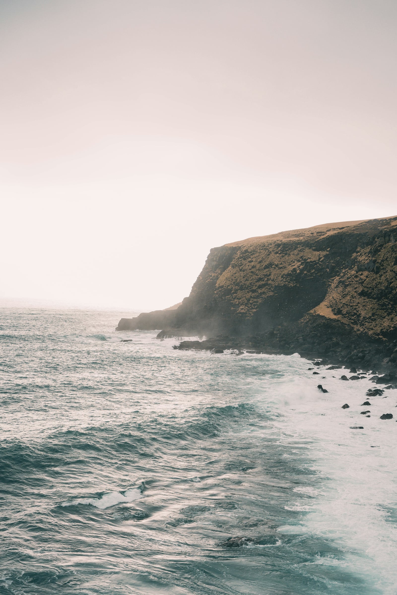 Soft ocean waves break below a grass-topped coastal cliff under a pale cream-gray sky.