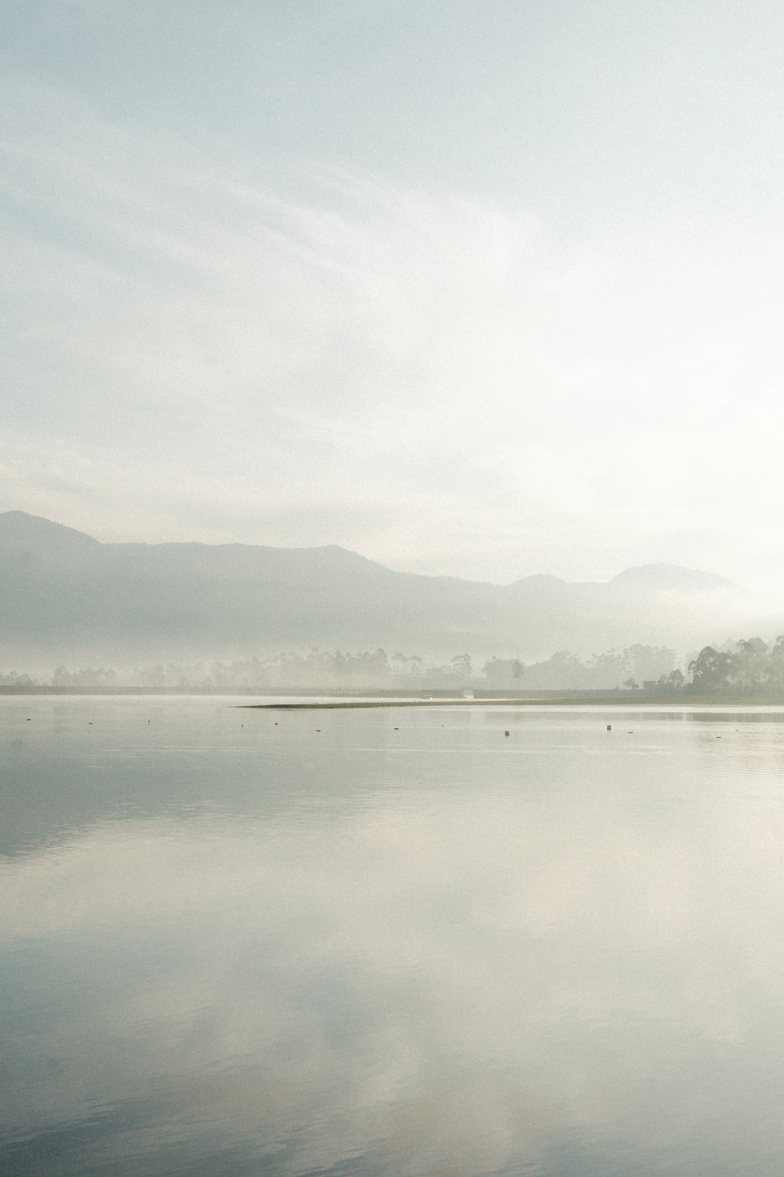 A still lake reflects a light sky while mountains and shoreline trees fade into morning mist.
