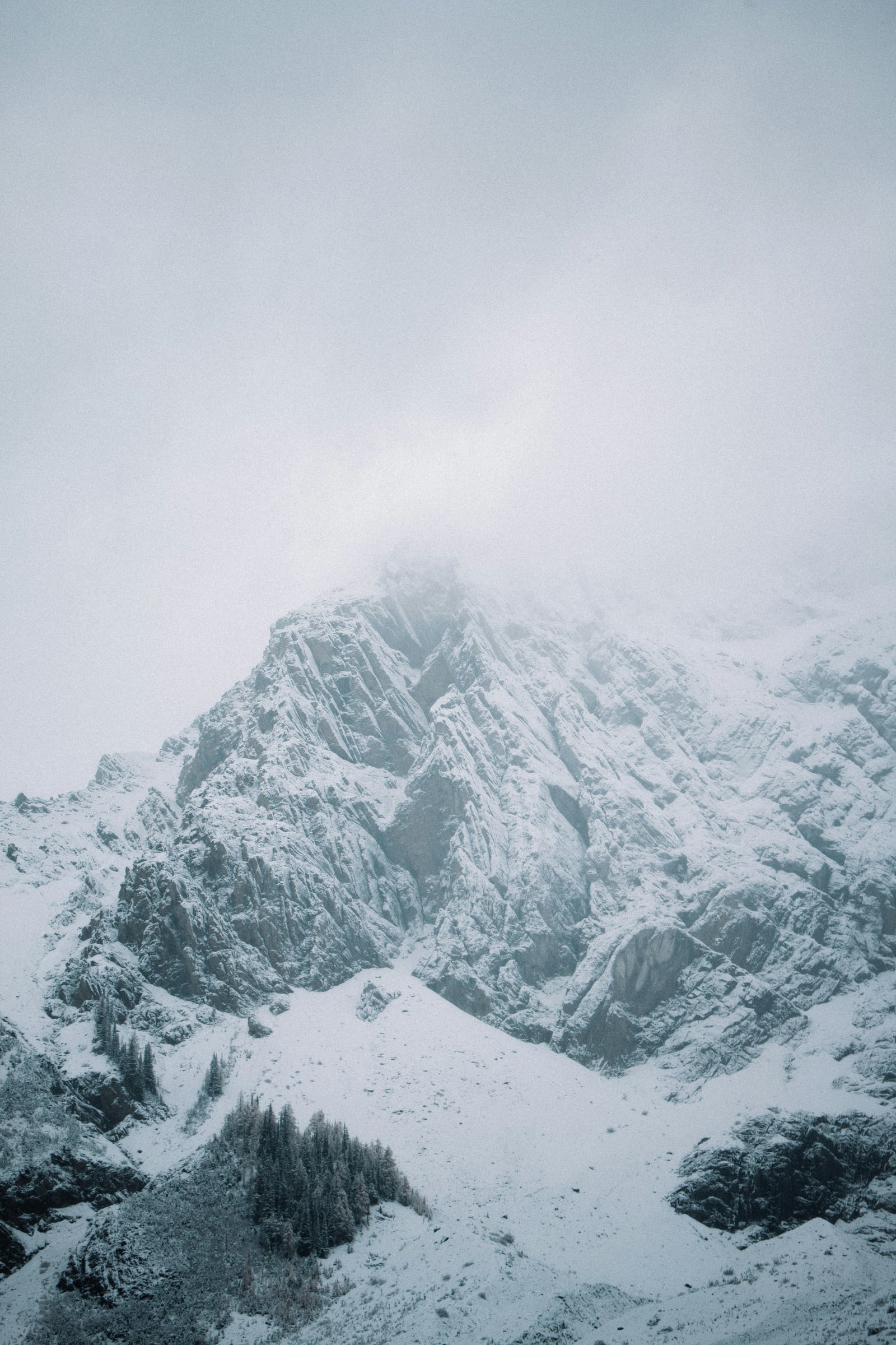 A snow-covered rocky mountain and small conifer groves sit beneath low pale gray clouds.