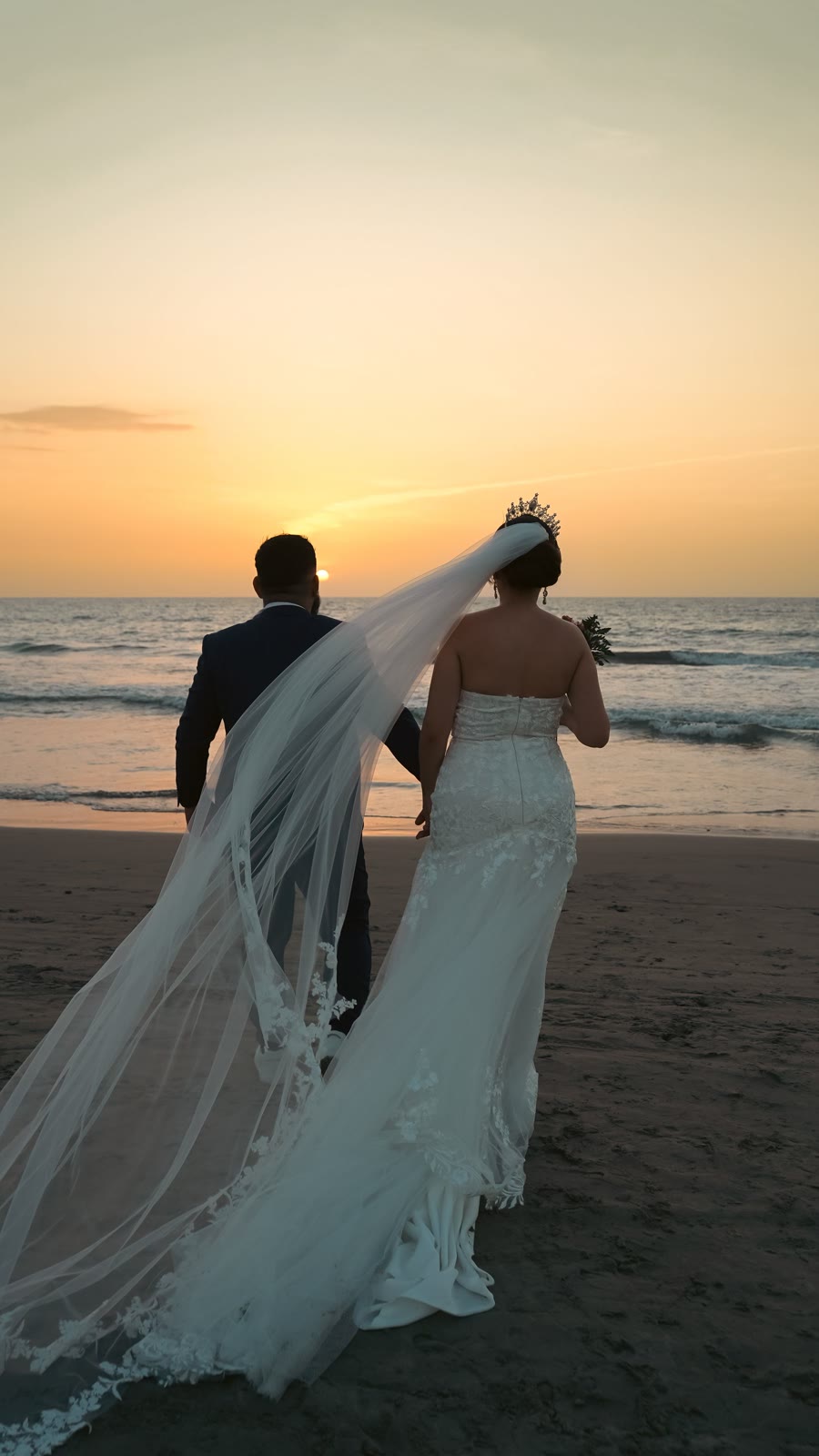 A couple in wedding attire walking beside the ocean at sunset.