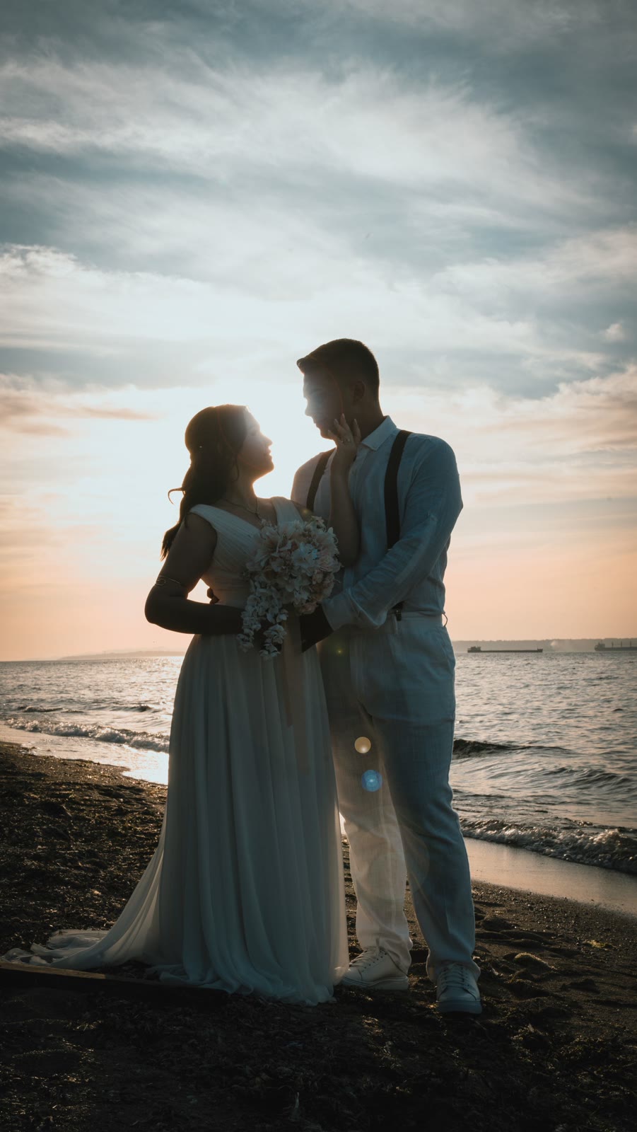 A newlywed couple embracing on a coastline in warm sunset light.